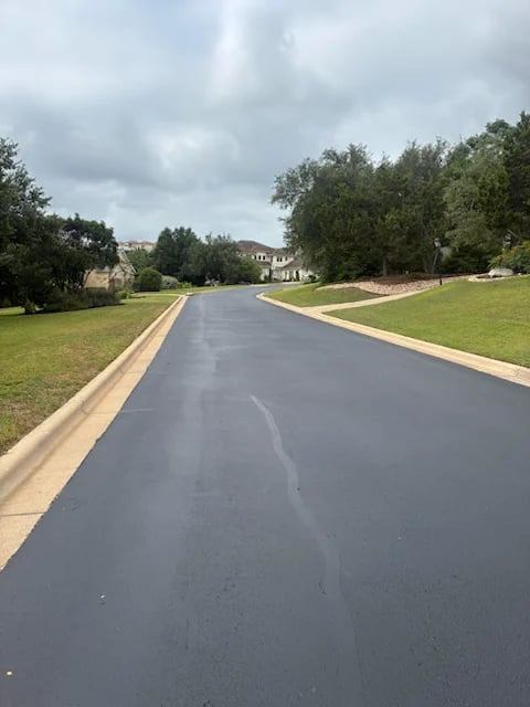 Paved driveway lined with manicured lawns and mature trees leading toward a residential home under cloudy sky — asphalt paving project in Central Texas by Austin Premier Paving