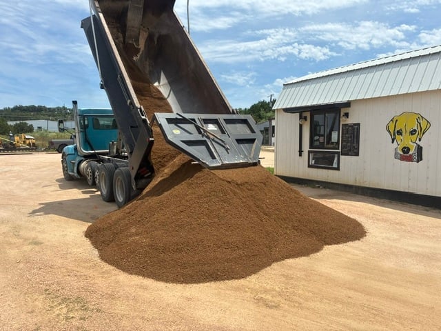 Dump truck unloading mulch onto sandy ground at a landscaping facility with office building visible