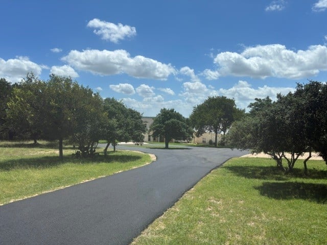 Winding asphalt driveway lined with mature oak trees and green lawns under a blue sky with scattered clouds