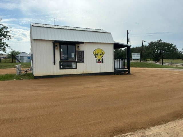 Small white metal building with yellow dog logo on rural dirt lot with wooden porch and surrounding fencing
