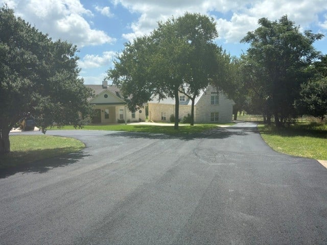 A large cream-colored house surrounded by mature trees with a curved asphalt driveway leading to the entrance