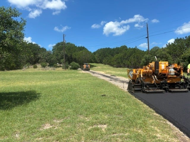 Yellow asphalt paving machine on a rural road with green grass fields and trees under blue sky