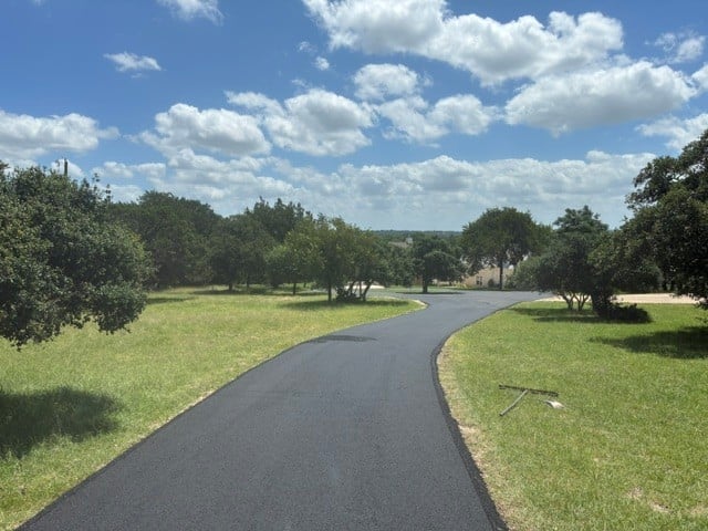 Winding paved path through green park with scattered trees and pond under blue sky with white clouds