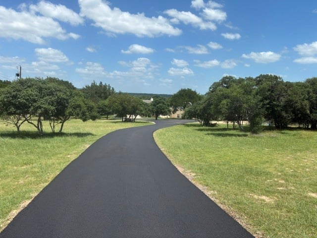 Winding black asphalt driveway curving through a grassy landscape with scattered trees under a blue sky with white clouds
