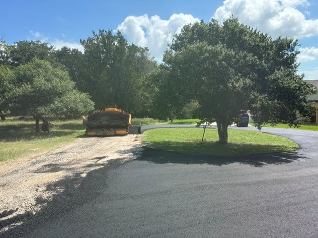 Yellow excavator on dirt driveway with mature trees and paved road under sunny blue sky